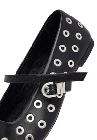 Scarpa piatta in pelle nera con occhielli decorativi in argento, cinturino alla caviglia regolabile e design della punta arrotondata. Texture liscia.