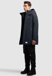 Marineblå vandtæt lang frakke med hætte, der har lynlås-lommer og en label ved kanten, parret med sorte cargobukser og støvler.