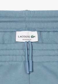 Kirkas siniset puuvillahousut, joissa on elastinen vyötärö, kaksi narua, ja Merkit "LACOSTE" sekä "REGULAR FIT", sekä teksturoitu ommel.