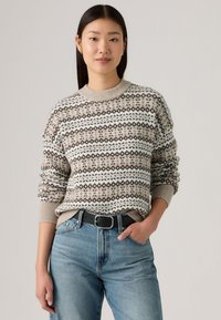 Gebreide trui met een beige, witte en bruine patroon en geometrische ontwerpen. Ribgebreide mouwen en kraag, gedragen met lichtblauwe jeans en een zwarte riem.