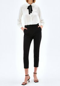 Vrouw in een witte blouse met zwarte strik, zwarte op maat gemaakte broek tot de enkels en zwarte sandalen met hoge hakken, staand met handen in de zakken.