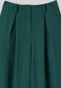 Pantalon taille haute vert foncé avec plis et passants de ceinture, fabriqué en tissu texturé.