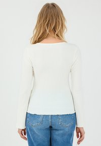 Maglietta a maniche lunghe a coste di colore bianco sporco con scollatura rotonda, design aderente e orlo a scallop. Abbinata a jeans a vita alta di colore blu chiaro.