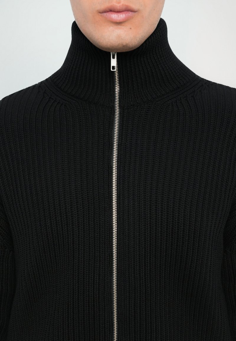 Pull en tricot noir avec un col haut et une fermeture éclair en argent sur toute la longueur, présentant une texture côtelée verticale et une silhouette ajustée.