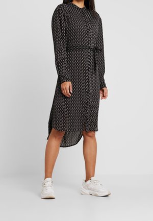 Femme portant une robe midi noire à manches longues avec motif, ceinture à la taille et baskets blanches, debout devant un fond gris uni.