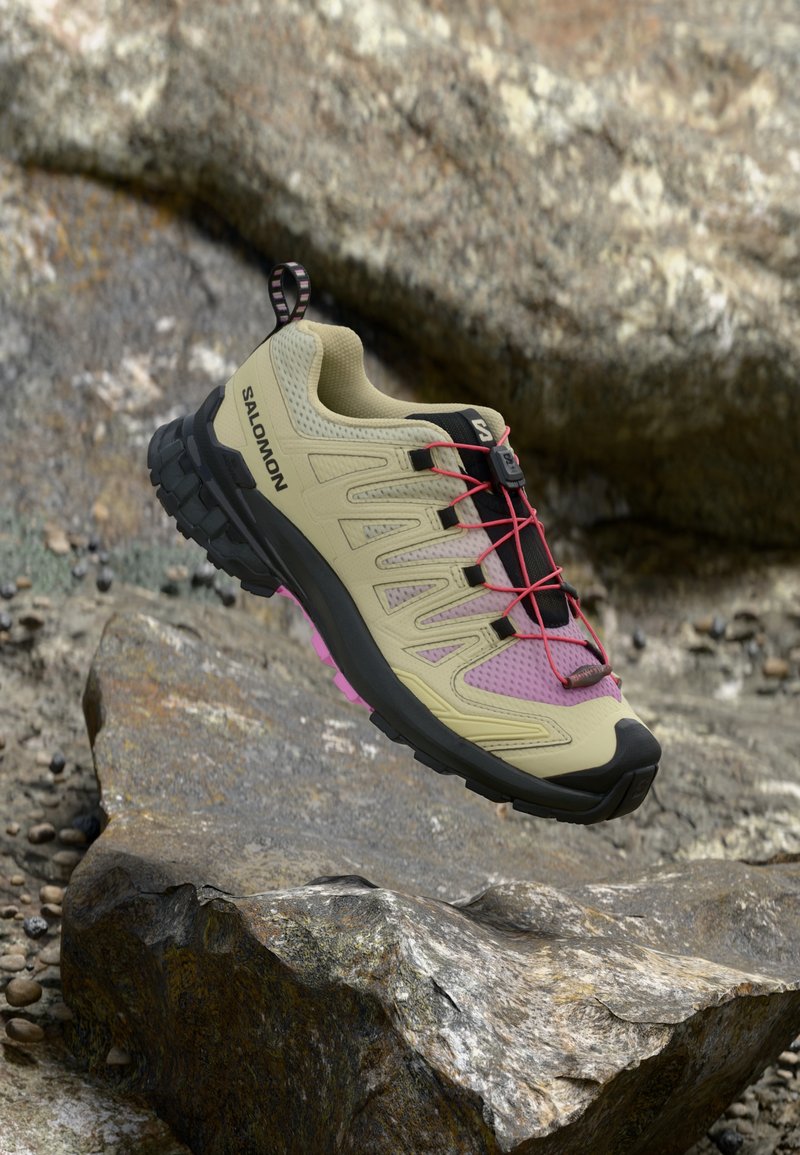 Scarpa da trail running beige con accenti rosa, pareti in rete testurizzata, suola in gomma nera e sistema di lacci rossi. Caratterizzata da una linguetta sul tallone.