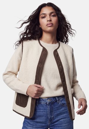 Femme aux longs cheveux bouclés porte un pull crème, une veste polaire crème et marron, et un jean bleu, affichant une assurance avec la main sur la veste.