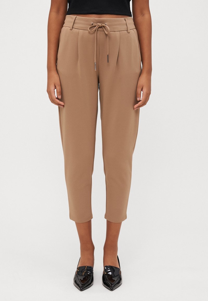 Pantalon court marron avec plis devant et taille à cordon, porté avec des chaussures vernies noires à bout pointu.