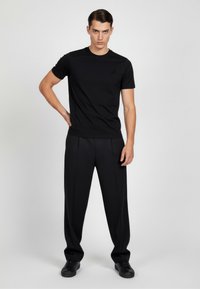 Camiseta negra de manga corta con un logo sutil, combinada con pantalones negros con pinzas y zapatillas negras, sobre un fondo neutral.