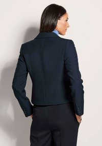 Navy blazer van gladde stof, met een getailleerde pasvorm, ingesneden revers en knopenaccenten op de mouwen. Achteraanzicht met een slanke uitstraling.