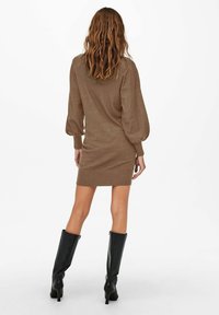 Robe en tricot marron avec de longues manches ballon, une silhouette ajustée et des poignets côtelés, associée à des bottes en cuir noires montant jusqu'aux genoux.