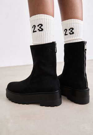 Piernas con calcetines blancos acanalados con "23" en negro y botines de ante negros con suelas gruesas y cremalleras traseras sobre un suelo claro.