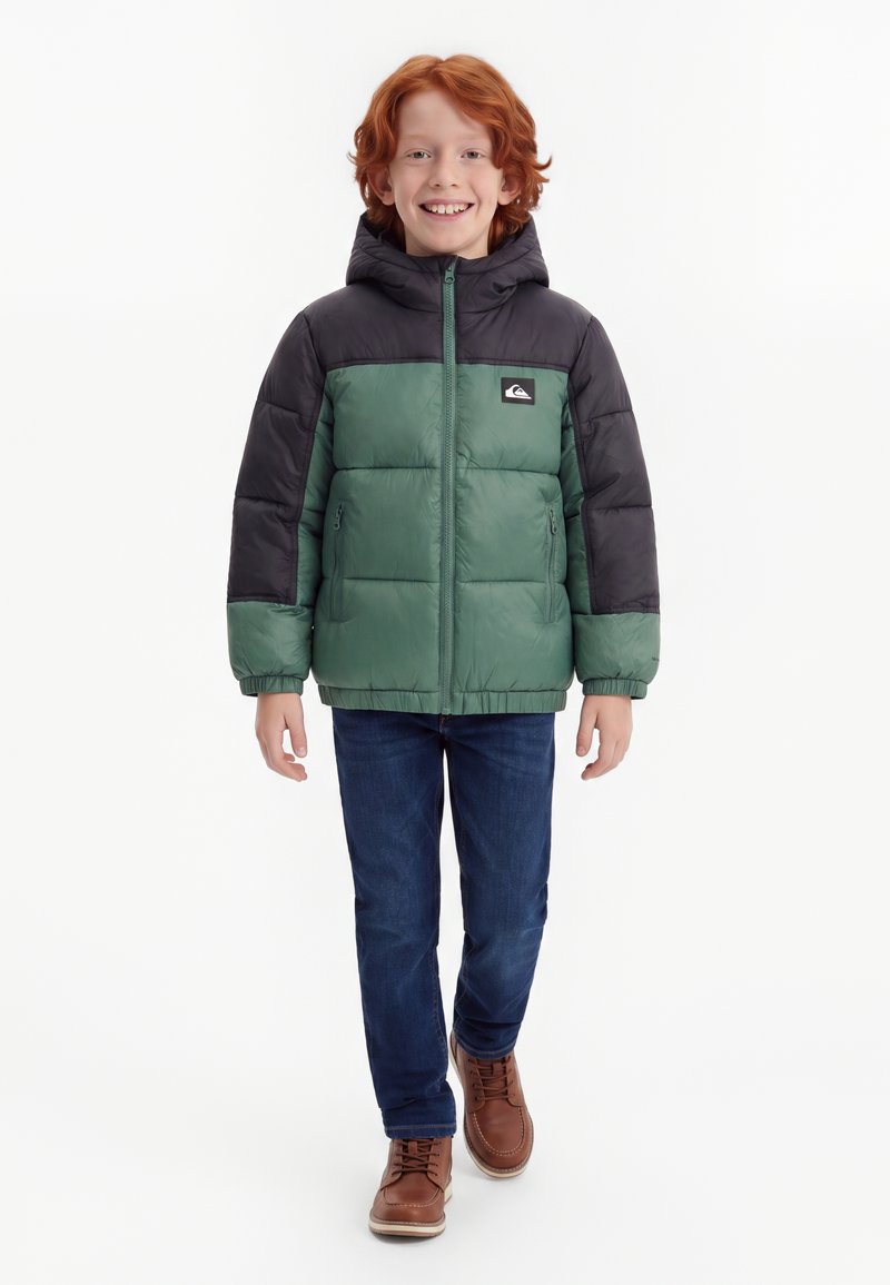 Enfant aux cheveux roux portant une veste à capuche verte et noire, un jean bleu et des chaussures marron, marchant vers l'avant et souriant.