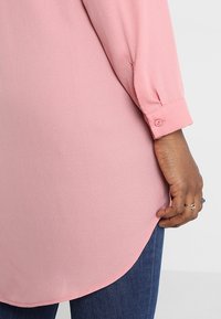 Blouse rose clair à texture lisse, manches longues, poignets boutonnés, et ourlet arrondi, associé à un jean bleu foncé.