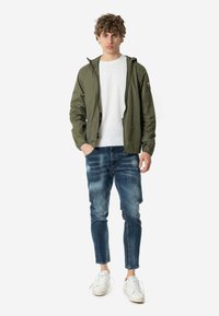 Chaqueta ligera verde con capucha, camisa blanca debajo, vaqueros azules deslavados y zapatillas blancas. Textura suave, ajuste casual y diseño sencillo.