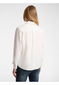 Camicia bianca a manica lunga con colletto, dettaglio con piega sul retro e polsini abbottonati, abbinata a jeans blu scuro. Texture liscia.