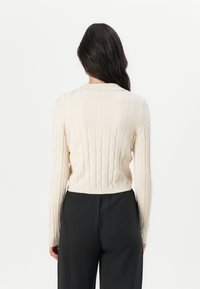 Beige ribgebreide trui met een ronde kraag, lange mouwen en een cropped ontwerp, met kabelbrei details langs de achterkant.