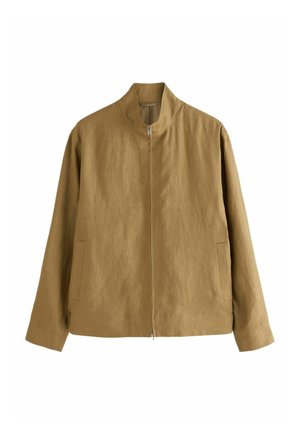 Man wearing tan zip-up jacket, beige cap, black pants, and black sandals standing near stone steps against white wall.