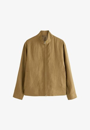 Man wearing tan zip-up jacket, beige cap, black pants, and black sandals standing near stone steps against white wall.