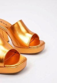 Mules de plataforma metálicos dorados con diseño de punta abierta, textura suave y tacón cuadrado. Parte superior redondeada con una amplia abertura.