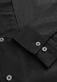 Black shirt made of smooth fabric, featuring a folded cuff with two gray buttons, showing fine stitching and a clean design.