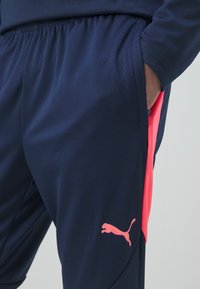 Marineblaue Sporthose mit strukturierter Oberfläche, ausgestattet mit einem leuchtend pinken seitlichen Streifen und einem pinken Puma-Logo. Elastischer Bund und Seitentaschen.