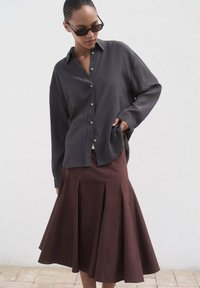 Camicia button-up grigia scura a maniche lunghe abbinata a una gonna marrone svasata con pieghe, indossata con occhiali da sole neri rettangolari.