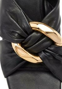 Sandale en cuir noir avec un design supérieur noué et un détail en métal doré. Texture lisse avec une semelle plate et sans talon.