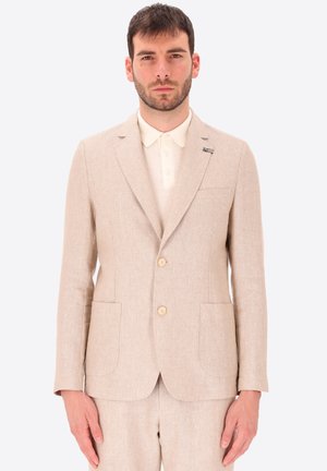 Uomo che indossa giacca beige a due bottoni e pantaloni coordinati sopra una camicia bianca con colletto, in piedi contro uno sfondo semplice.