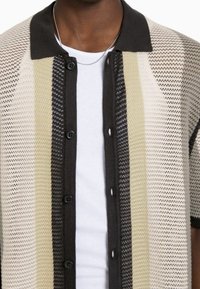 Camisa de botón de manga corta con un diseño de malla de punto que presenta rayas alternas en colores beige, negro y crema. Botones negros.