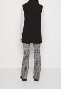 Svart ärmlös väst med krage, kombinerad med grå och vit mönstrad utsvängd byxor. Slät textur, minimalistisk design, mångsidig outfit.