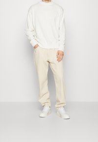 Felpa bianca a maniche lunghe con scollo rotondo, abbinata a pantaloni beige chiaro larghi e sneakers bianche, creando un outfit casual.