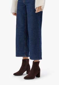 Botines de ante marrón con un tacón grueso, puntera afilada y cremallera lateral, combinados con jeans de mezclilla oscuros de pierna ancha.