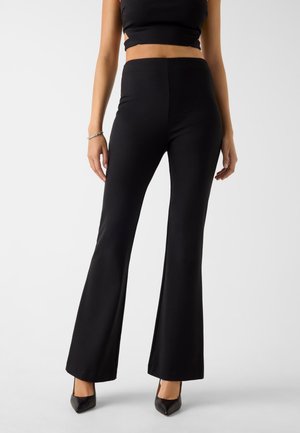 Pantaloni neri a zampa realizzati in tessuto elasticizzato, con vita alta e trama liscia. Abbinati a décolleté nere appuntite per creare contrasto.