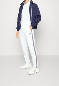 Námořnický větrovka s bílými a červenými akcenty přes bílý polo triko, doplněná bílými sportovními kalhotami s postranními pruhy a logem, a bílými teniskami.