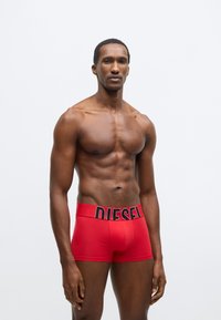 Červené boxerky z hladkej tkaniny s širokým čiernym pásom s logom "DIESEL". Majú priliehavý strih a dĺžku do polovice stehna.