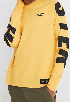 Chemise jaune à manches longues avec le texte noir "Hollister" sur les manches, logo noir sur la poitrine, et un patch de marque noir près de l'ourlet, portée avec un pantalon vert.