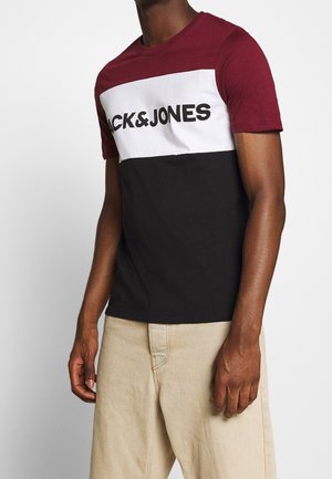 Homme portant un t-shirt color-block en bordeaux, blanc avec un texte logo noir, associé à un pantalon beige, sur un fond uni.