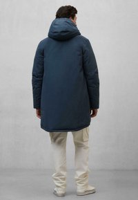 Ciemnoniebieska kurtka puchowa z kapturem, szwami na ramionach i rozcięciem na plecach, noszona na jasnobeżowych spodniach cargo i białych sneakersach.