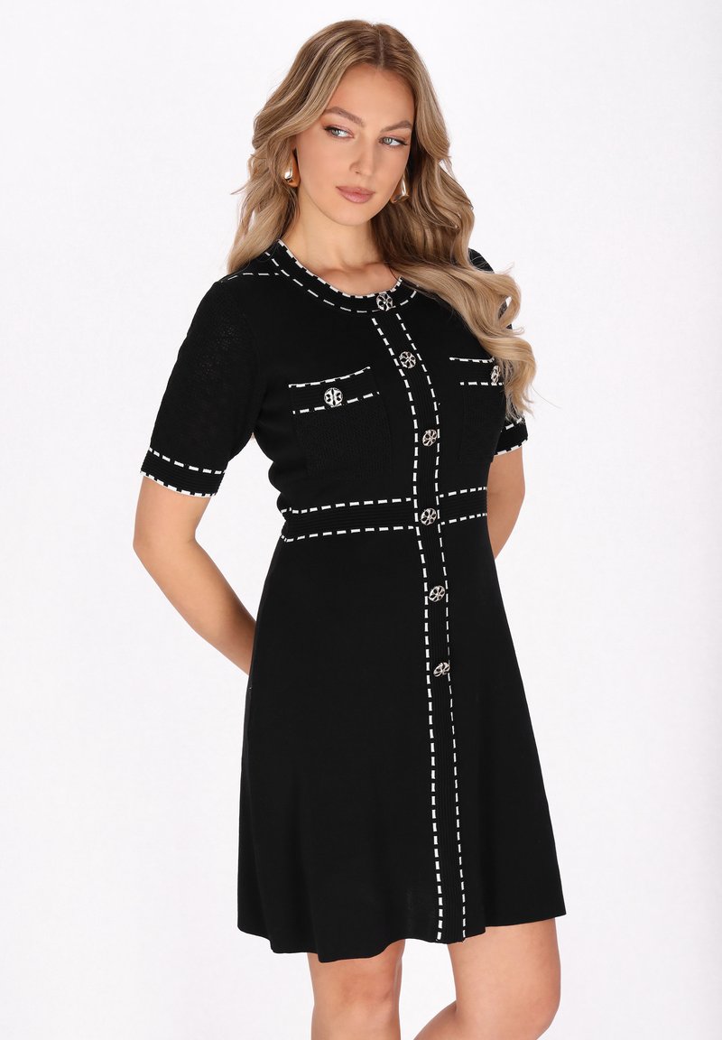 Abito nero realizzato in tessuto a maglia, con linee bianche cucite, due tasche frontali e bottoni decorativi. Maniche corte, silhouette a linea ad A.