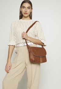 Borsa a tracolla in pelle marrone con pattina arrotondata, chiusura con zip e dettaglio a frange. Texture liscia, cuciture sottili e tracolla regolabile.