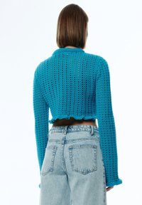 Gekürzter, gestrickter Pullover in leuchtendem Türkis mit strukturiertem, offenem Gewebe, langen Ärmeln und rüschengesäumtem Saum, kombiniert mit hellblauen Jeans.