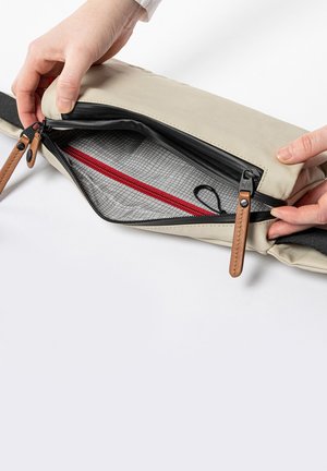 Hände halten eine offene beige Bauchtasche, die einen schwarzen Reißverschluss und ein graues Innenfutter mit einer roten Reißverschlusstasche innen zeigt.