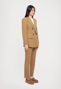 Kvinne står iført en beige dobbeltradet blazer med matchende bukser og brune platåloafers mot en ensfarget hvit bakgrunn.