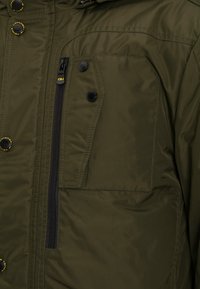Olivgrüne gepolsterte Jacke mit einer Reißverschlusstasche auf der Brust, Druckknöpfen und strukturiertem Stoff. Merkmale sind kontrastierende schwarze Reißverschlüsse und Akzente.