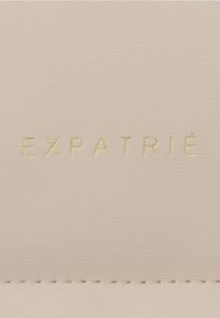 Pelle beige testurizzata con la stampa in rilievo "EXPATRIÉ" in oro. Superficie liscia con dettagli di cucitura sottili alla base.