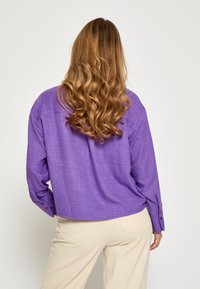 Lilla linned skjorte med løs pasform, lange ærmer, rundet bottom og knapdetaljer ved manchetterne. Bæres med lysfarvede bukser.