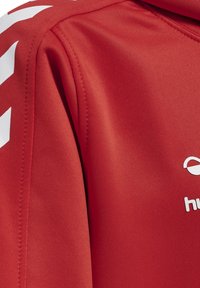 Giacca sportiva rossa con una texture liscia, caratterizzata da accenti angolari bianchi sulle spalle e un logo stampato sul petto.