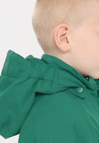 Grøn polstret jakke med hætte, der har et samlet design og trykknapper. Teksturindbundet stof med synlige syningsdetaljer.