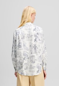 Camicia bianca con stampa di un caffè e un edificio disegnati a linee, maniche lunghe e fondo curvo. Il tessuto ha una consistenza liscia e leggera.